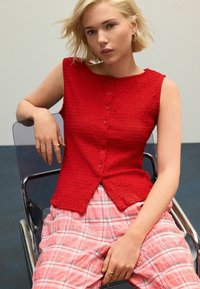 Haut sans manches texturé rouge avec un plastron de boutons, associé à un pantalon plissé en vichy rose. Modèle assis sur une chaise transparente.