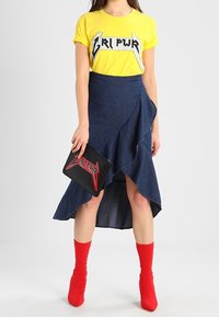 Donna che indossa una maglietta gialla con la scritta "GRL PWR", gonna asimmetrica blu scuro con volant, stivaletti rossi alla caviglia e tiene in mano una clutch nera con la stessa scritta "GRL PWR".