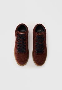 Brown suede sneakers with dark blue laces, perforated accents, and a gum outsole. Brand logo visible on the tongue.
