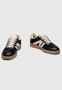 Le sneakers nere e crema presentano una tomaia in suede e pelle, lacci sottili e una suola in gomma naturale, il tutto con un design minimalista.