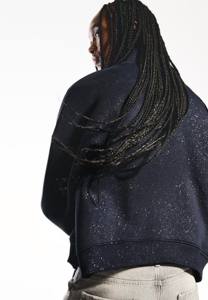 Sweatshirt blu navy con una vestibilità rilassata, caratterizzato da una texture maculata e orlo a coste. Completato da lunghi capelli intrecciati visibili sullo sfondo.