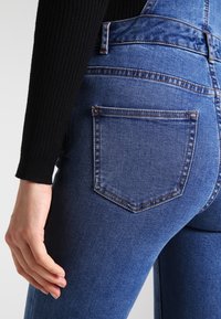 Denimoveraller i en medelblå tvätt. Har en hög midja, figursydd silhuett och bakfickor med en klassisk rak skärning.