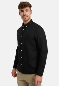 Mladý muž s kučeravými vlasmi, oblečený v čiernej košeli na gombíky a béžových nohaviciach, stojaci pred jednoduchým svetlým pozadím.