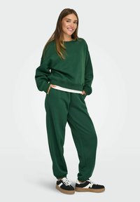 Grøn sweatshirt og matchende bukser lavet af blødt stof. Bukserne har elastiske manchetter og lommer. Sneakers har sorte, hvide og brune detaljer.