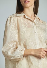 Blusa ricamata beige chiaro con colletto, caratterizzata da motivi floreali, chiusura frontale con bottoni e polsini elasticizzati, realizzata in tessuto trasparente.
