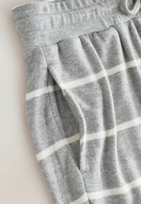 Pantaloni de trening gri deschis cu dungi albe orizontale, având un bandou elastic la talie și o buzunar lateral.