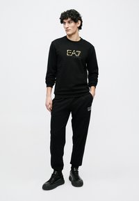 Musta collegepaita kultaisella logolla, mustat verkkarit ja mustat lenkkarit. Sileä kangas, rento design, jossa logo-yksityiskohdat edessä ja housuissa.