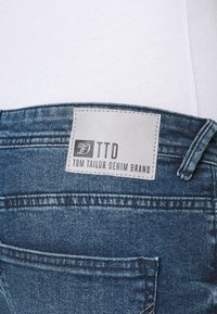 Jeans i denim med en medelblå tvätt, med en läderliknande patch märkt "TTD" och "TOM TAILOR DENIM BRAND" nära midjebandet.