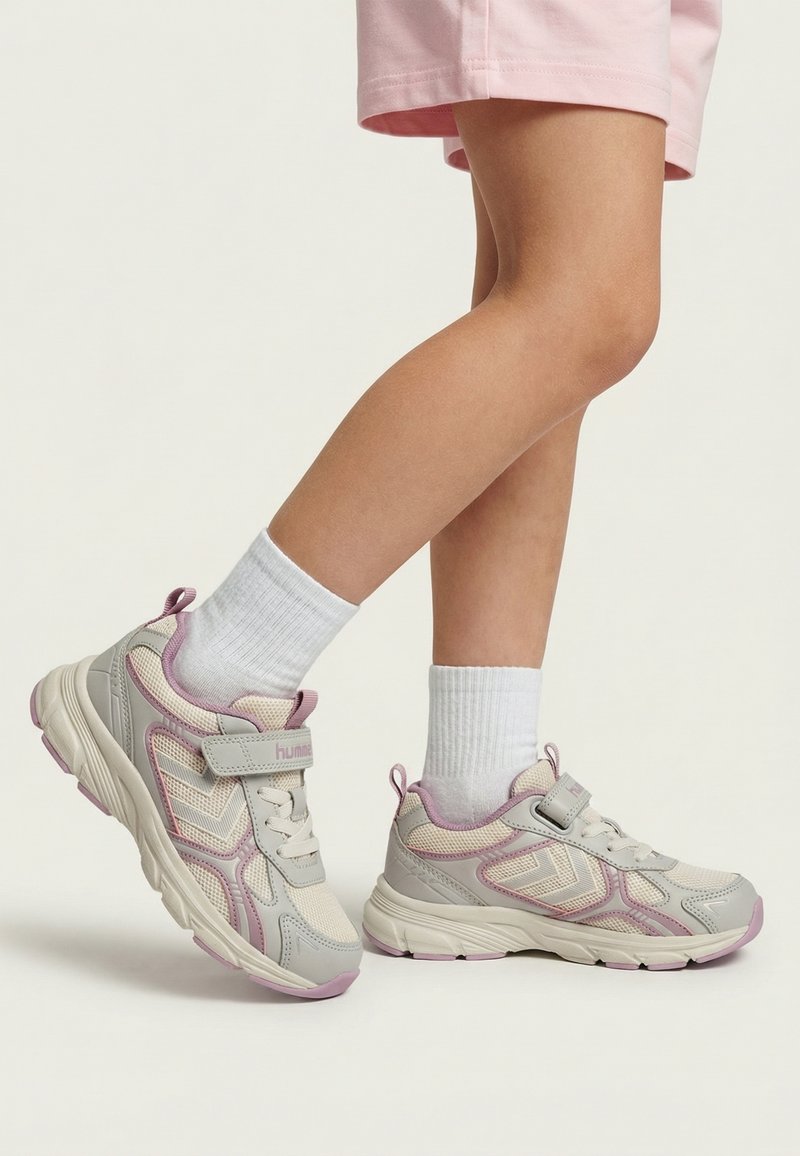Kind draagt witte en roze sportsneakers met witte enkelsokken en roze korte broek, staand op een effen lichte achtergrond.