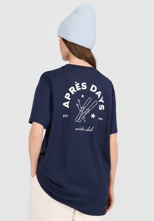Osoba nosząca granatowy T-shirt z napisem "APRÈS DAYS" i grafiką skrzyżowanych nart na plecach, jasnoniebieską dzianą czapkę i jasne spodnie.