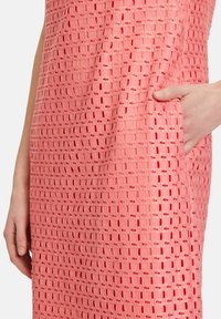 Rosa strukturiertes Kleid mit einem gewebten Muster, kurzen Ärmeln und Seiten Taschen. Der Stoff weist sichtbare Ausschnitte und ein strukturiertes Design auf.