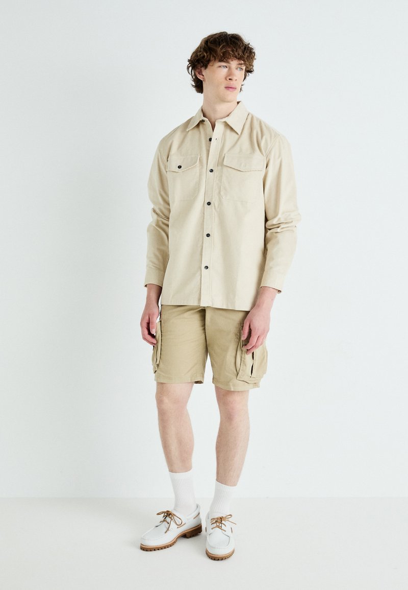 Chemise beige à manches longues avec deux poches poitrines et boutons noirs, accompagnée de shorts cargo beiges et de chaussures blanches avec des semelles brunes.