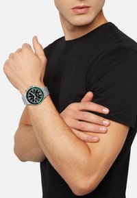 Sølvmetal ur med en sort urskive, grøn bezel og tre underdials, båret på et håndled med lys hudtone, iført en sort t-shirt.