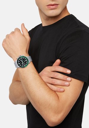 Montre en métal argenté avec un cadran noir, une lunette verte et trois sous-cadrans, portée sur un poignet au teint clair, avec un t-shirt noir.