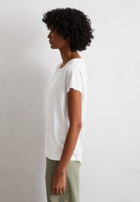 Witte T-shirt met korte mouwen, een relaxte pasvorm, rond decolleté en subtiele details op de borst; gecombineerd met lichtgroene broek.