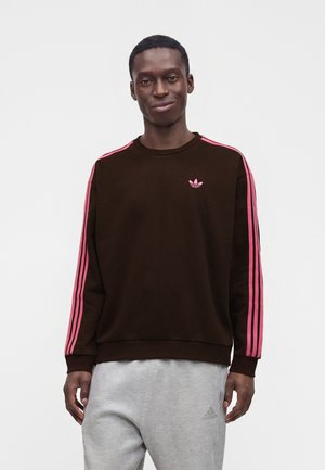 Άνδρας που φοράει μαύρη μπλούζα Adidas με ροζ ρίγες στα μανίκια και γκρι φόρμα Adidas στέκεται μπροστά σε ένα απλό λευκό φόντο.