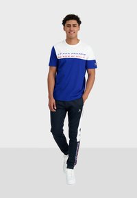 T-shirt à manches courtes bleu et blanc avec l'imprimé "LE COQ SPORTIF", associé à un jogging noir avec des panneaux latéraux blancs et un logo. Sneakers blanches.