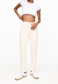 Jean taille haute beige clair avec une coupe droite, dotés de poches avant et de coutures discrètes, portés avec un t-shirt court blanc et des baskets.