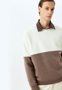 Suéter de cuello redondo con un diseño bicolor en secciones blanca y marrón. Tejido suave, cuello y puños de canalé con un sutil detalle de logo.
