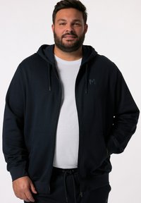 Felpa navy con cerniera, tasche a canguro, cappuccio con cordino e piccolo logo. Tessuto morbido con una superficie liscia. Abbinata a una t-shirt bianca.