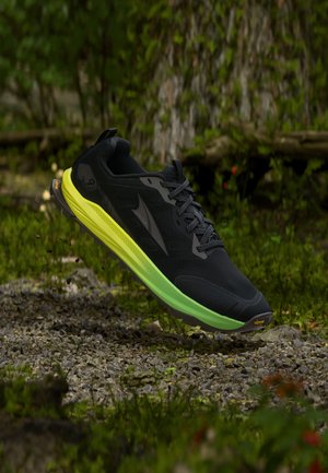 Chaussure de trail running noire et verte en l’air au-dessus d’un chemin de gravier avec feuillage vert et tronc d’arbre en arrière-plan.