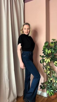 Γυναίκα με ξανθά μαλλιά που φοράει μαύρο τοπ και σκούρα μπλε τζιν πόζάρει δίπλα σε μια γλάστρα με πράσινα φύλλα, μπροστά σε ροζ τοίχο και μπεζ κουρτίνα.