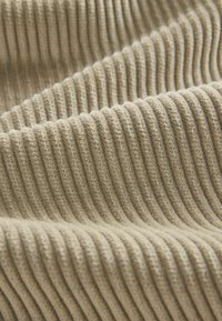 Beige värjätty kangas, jossa on pehmeä rakenne ja tasaisesti sijoitetut pystysuuntaiset viivat, muodostamassa hienovaraista aaltokuviota pinnalle.