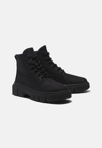 Bottines noires à lacets avec semelles épaisses, massives et une bande de roulement texturée sur un fond clair uni.