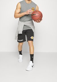 Szary bezrękawnik, czarne spodenki z napisem "LAKERS"; szara bluza zawiązana wokół talii; białe trampki; trzyma piłkę do koszykówki.