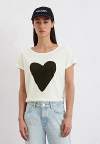 Lichtgekleurde t-shirt met een grote zwarte hartafbeelding op de voorkant, korte mouwen, ronde halslijn, gecombineerd met lichtblauwe jeans.