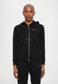 Crna zip majica od mekanog pamučnog materijala, s kapuljačom, prednjim džepovima i zlatnim logotipom na prsima. Slim fit dizajn s rebrastim otvaranjima na rukavima.