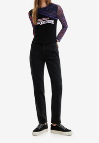 Schwarzes Langarmoberteil mit lila Netzärmeln und dem Text "Happige Zeiten kommen", kombiniert mit schwarzen Jeans mit Verzierungen und schwarzen Turnschuhen.