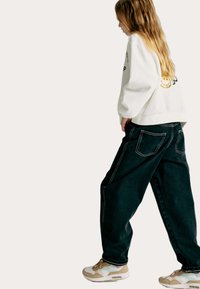 Sweat-shirt blanc surdimensionné avec impression graphique, jean baggy vert foncé, et baskets beiges avec des accents pastel. Vue de côté, tenue décontractée.