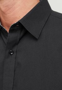 Svart skjorta med knappar och strukturerad krage, med en subtil textur och kontrasterande detalj på insidan av kragen. En svart knapp är synlig.