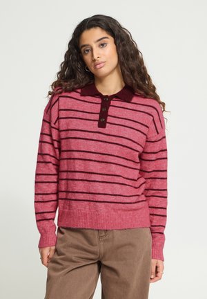 Jeune femme aux longs cheveux bouclés portant un pull rayé rose et rouge foncé ainsi qu’un pantalon marron, debout devant un fond uni.