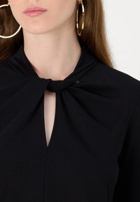 Svart topp med en front nøkkelhulldesign og en tvunnet knut ved utringningen, laget av et glatt stoff. Store gull hoopsølv.