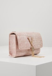 Růžová kabelka z umělé krokodýlí kůže s plochým klopovým zapínáním, zlatými kovovými prvky a řetízkem s třásněmi; má kompaktní obdélníkový tvar.