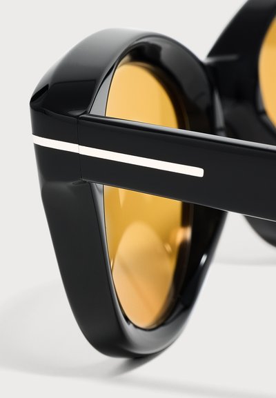 Gros plan sur des lunettes de soleil noires avec des montures épaisses et des verres teintés ambre, montrant un détail d'une bande blanche sur la branche.