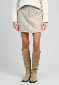 Jupe beige hauteur genou avec des embellissements argentés dispersés, associée à des bottes en suède taupe hautes. Texture douce et coupe ajustée.