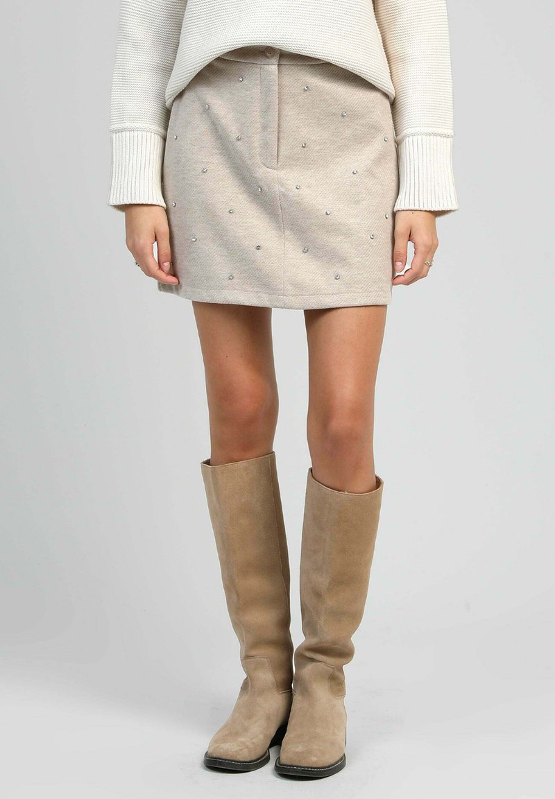 Jupe beige hauteur genou avec des embellissements argentés dispersés, associée à des bottes en suède taupe hautes. Texture douce et coupe ajustée.