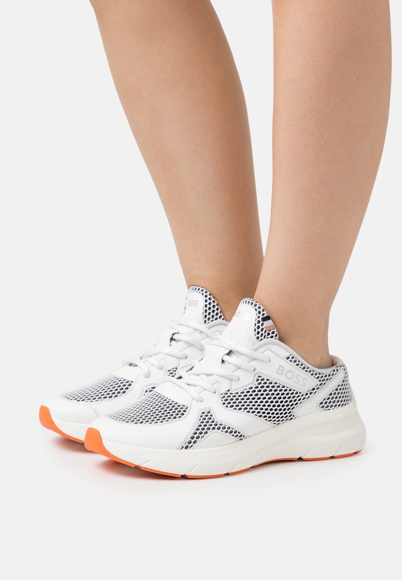 Personne portant des baskets de sport blanches avec des détails en mesh noir et des semelles orange, pieds joints sur un fond clair uni.