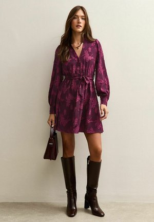 Pruimenkleurige jurk met een ceintuur in de taille, lange mouwen en bloemenstructuur; gecombineerd met bruine kniehoge laarzen en een mini handtas.