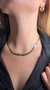 Κολιέ σε χρυσή απόχρωση με σχεδιασμό πλεκτής αλυσίδας και κεντρικό κούμπωμα. Συνδυασμένο με χρυσούς κρίκους σκουλαρίκια. Το φόντο απεικονίζει μαύρο πουκάμισο.