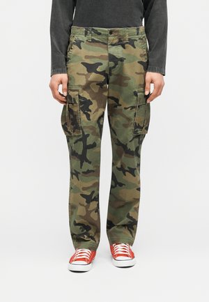 Uomo che indossa pantaloni cargo mimetici verdi e marroni, maglia nera a maniche lunghe e scarpe da ginnastica arancioni vivaci con lacci bianchi.