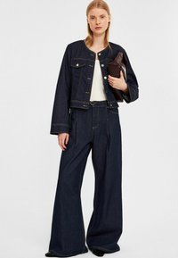 Mörk denimjacka med tryckknappar och framfickor, kombinerad med vida jeans med veck, bärs tillsammans med en vit topp och en brun clutch.