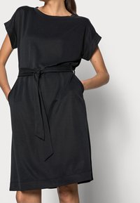 Schwarzes Kleid aus weichem Stoff, mit kurzen Ärmeln, einem Taillengürtel und Taschen, in gerader Schnittform und knielang.