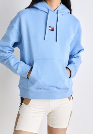 Osoba ubrana w jasnoniebieską bluzę z kapturem Tommy Jeans, z rękami w przedniej kieszeni, oraz kremowe szorty z brązowymi i czarnymi akcentami.