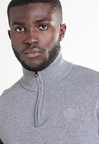 Szary dziergany sweter z wysokim kołnierzem i półzamek, z teksturowanym wzorem oraz małą naszywką z logo na piersi.