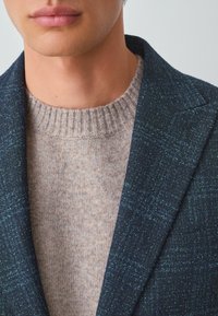 Marinblå jacka med textur och gröna detaljer, i kombination med en ljusbeige stickad tröja med ribbad halsringning.