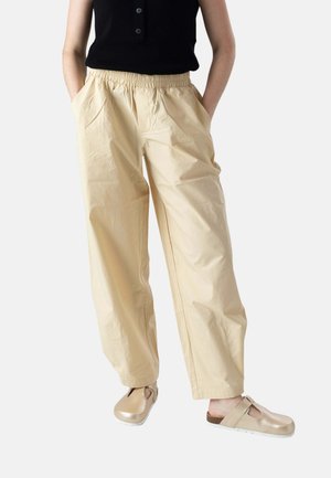 Persona con pantalones beige de pierna ancha con cintura elástica, camiseta negra sin mangas y zuecos beige de poner y quitar, de pie con las manos en los bolsillos.